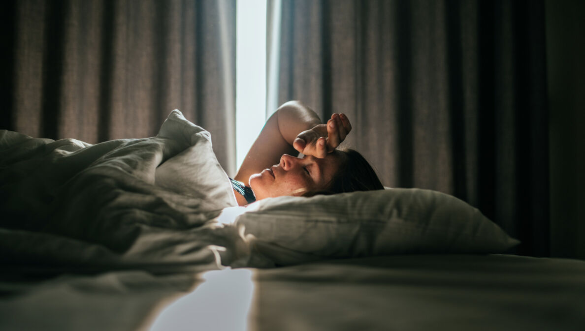 Eine Person liegt auf einem Bett, mit einem weißen Bettlaken und Kissen. Licht strömt durch ein Fenster, das von Vorhängen umgeben ist.