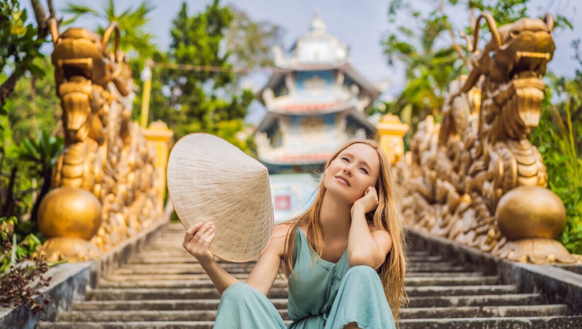 Eine Frau sitzt lächelnd auf einer Treppe. Sie schwenkt einen traditionellen vietnamesischen Hut. Im Hintergrund sind ein Tempel und goldene Statuen zu sehen.
