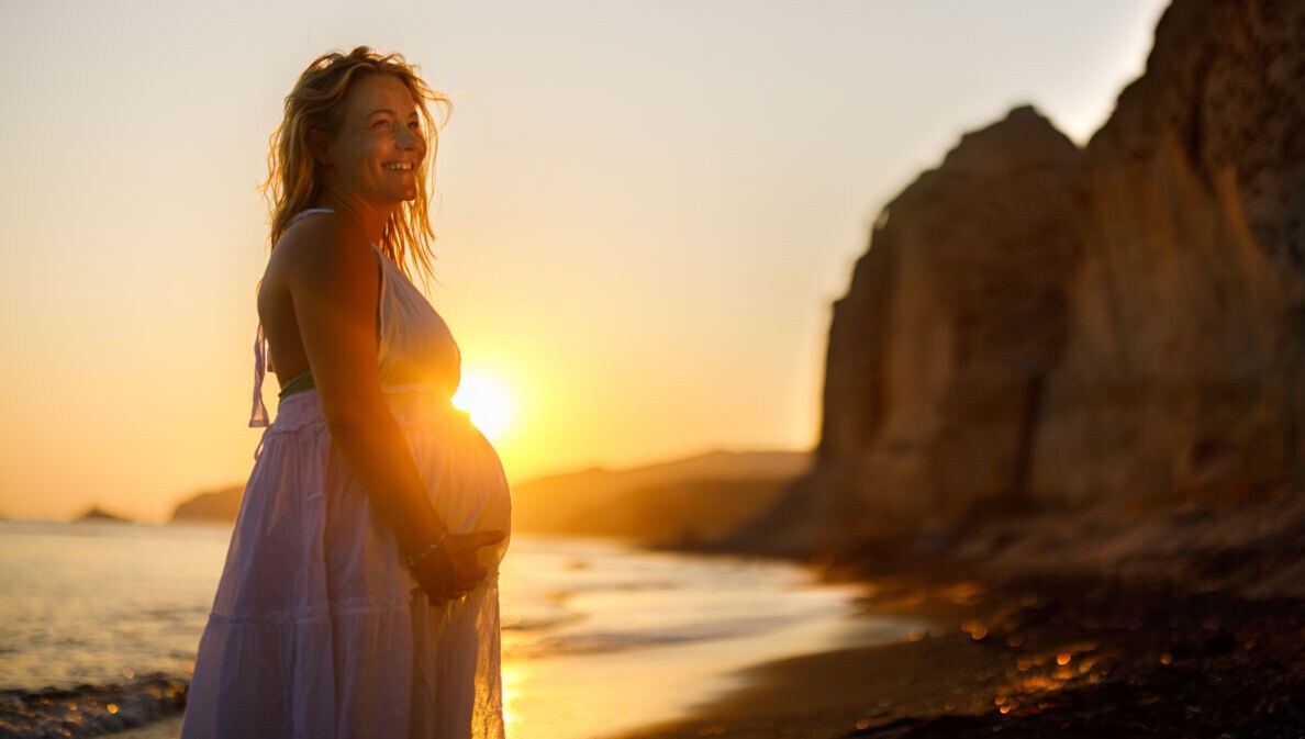 Eine lächelnde schwangere Frau im weißen Sommerkleid hält sich ihren Bauch an einem Strand mit Felsen bei Sonnenuntergang.