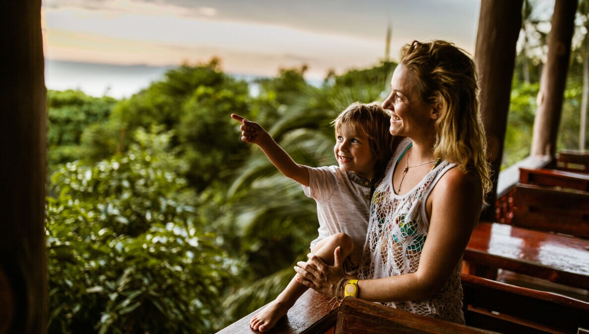 Eine Frau hat einen kleinen Jungen im Arm, beide blicken von einer Holzterrasse in den Dschungel von Thailand, hinter ihnen das Meer.