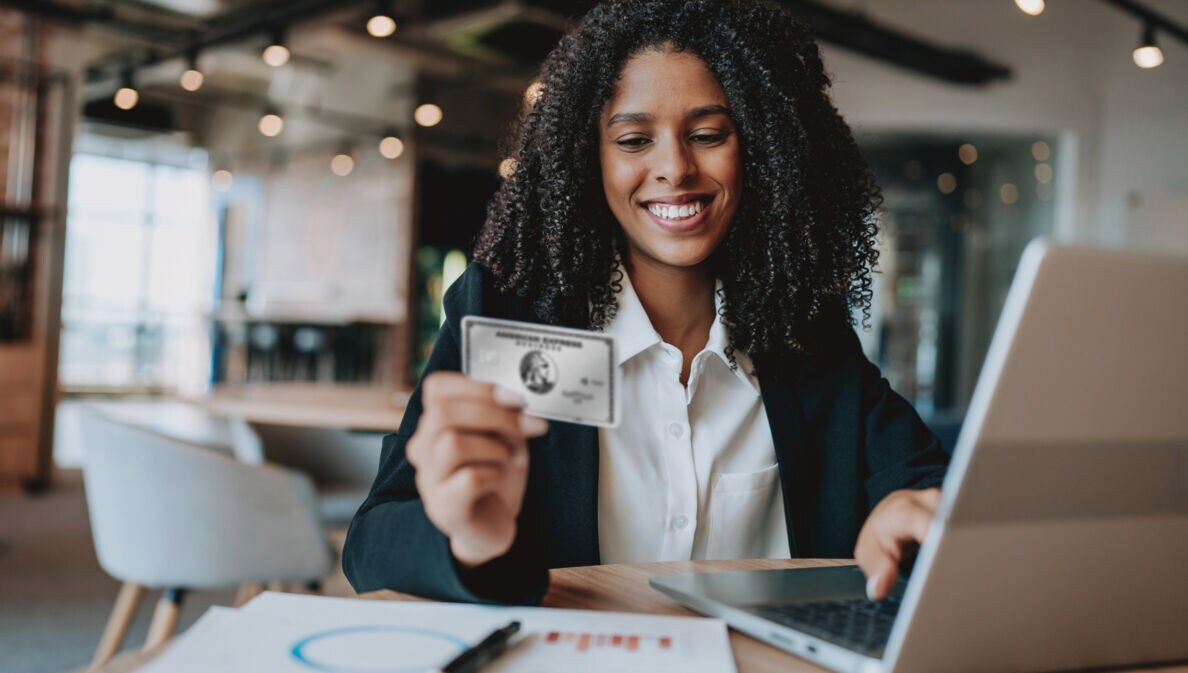 Eine lächelnde Frau bedient mit einer Hand einen Laptop und hält in der anderen Hand eine silberne Kreditkarte von American Express.