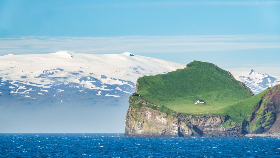 Blick auf die grüne isländische Insel Ellidaey mit einer einsamen Jagdhütte darauf und dem, unter einem Gletscher liegenden Vulkan Eyjafjallajökull im Hintergrund.