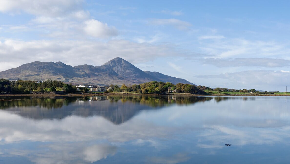 Vor einem großen Gewässer stehen kleine Häuser und Bäume, in Hintergrund ragt ein großer Berg empor, die Szene spiegelt sich in der Wasseroberfläche.