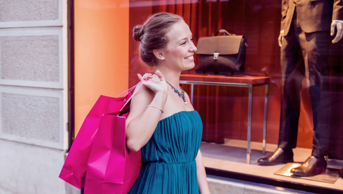 Eine lächelnde Frau im Kleid steht vor dem Schaufenster einer Modeboutique und hält zwei pinke Einkaufstüten über ihrer Schulter.