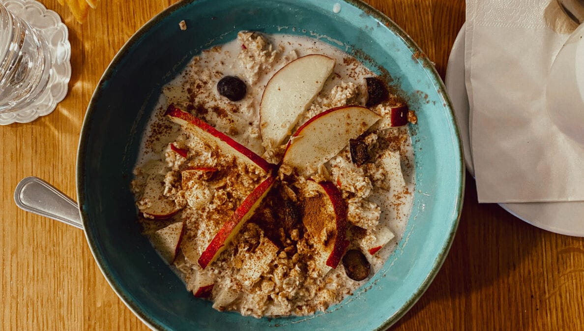 Blaue Schüssel mit Porridge, garniert mit Apfelscheiben, Blaubeeren und Zimt, auf Holztisch mit Glas Wasser und Serviette.