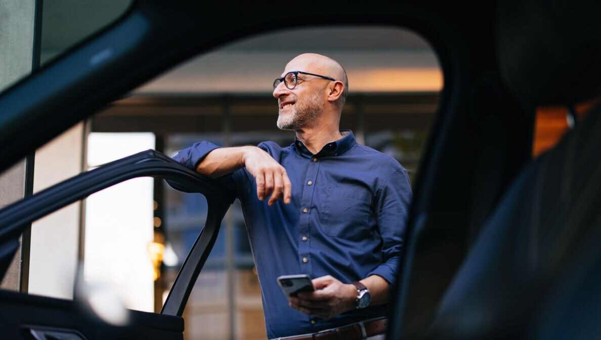 Ein Mann im blauen Hemd und mit einem Smartphone in der Hand lehnt an einer geöffneten Autotür und blickt lächelnd in die Ferne.