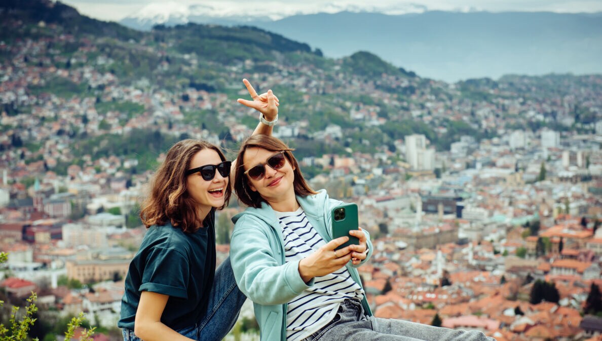 Zwei junge Frauen sitzen auf einer Mauer mit Blick auf eine Stadt, eine macht ein Selfie mit einem Smartphone und zeigt ein Peace-Zeichen.