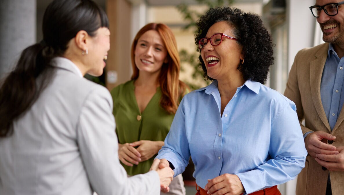 Vier Personen stehen beieinander in einem Büro, im Vordergrund geben sich zwei lachende Frau die Hand.