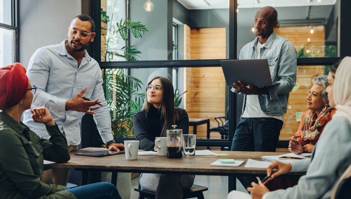 Mehrere Personen sitzen oder stehen an einem Tisch und schauen eine Person an, die spricht.