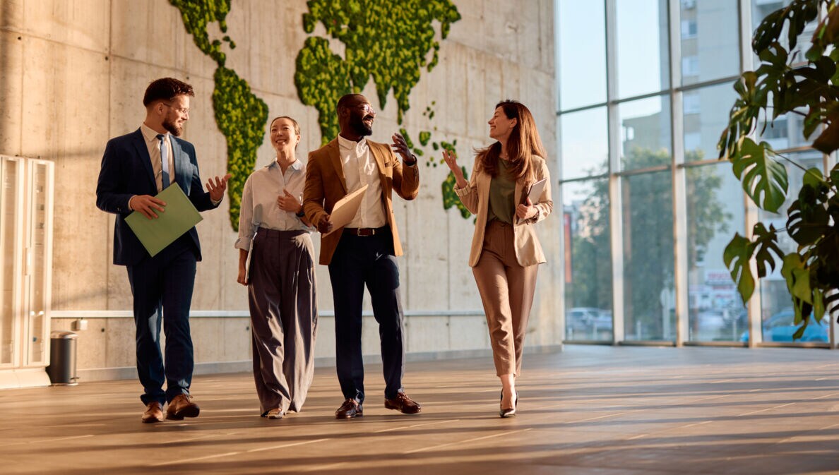 Vier Personen in Business-Outfits kommunizieren nebeneinander gehend in einem Foyer.