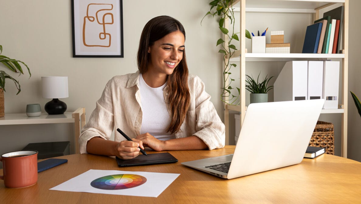 Eine lächelnde Frau arbeitet mit einem Grafiktablett an einem Laptop an einem Schreibtisch in einem Arbeitszimmer.