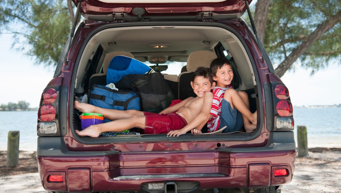 Zwei Jungen in Strandoutfits sitzen fröhlich in einem offenen Kofferraum, hinter ihnen das Meer.
