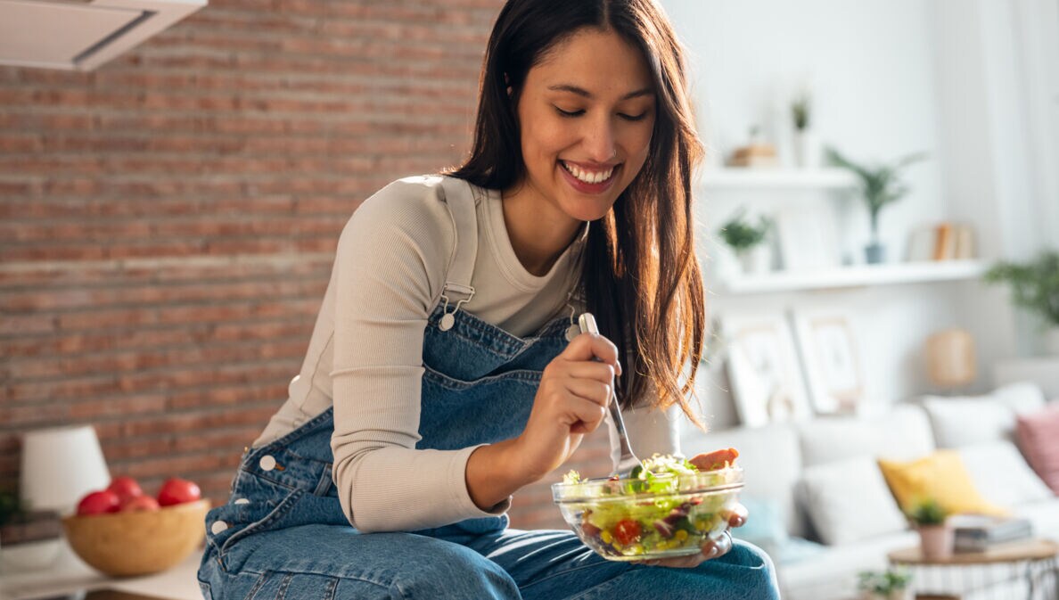 Frau in Jeanslatzhose sitzt auf einem Tisch und isst einen Salat aus einer Glasschüssel mit einer Gabel.