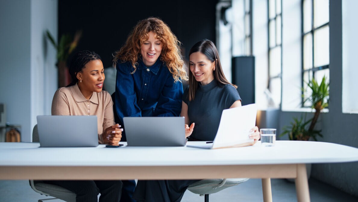 Drei Frauen schauen gemeinsam auf ein Laptop auf einem großen Tisch mit zwei weiteren Laptops in einem modernen Büro.