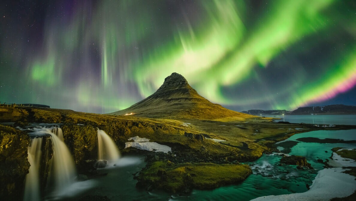 Moosbedeckte Vulkanlandschaft mit Wasserfällen unter Nachthimmel mit grün und lila leuchtenden Polarlichtern.