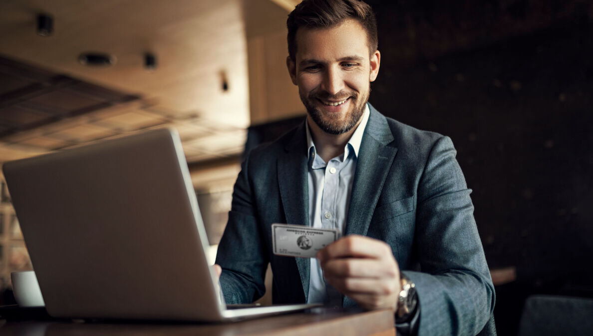 Ein lächelnder Geschäftsmann sitzt an einem Tisch vor einem Laptop und blickt auf eine silberne Platinum Kreditkarte von American Express in seiner Hand.