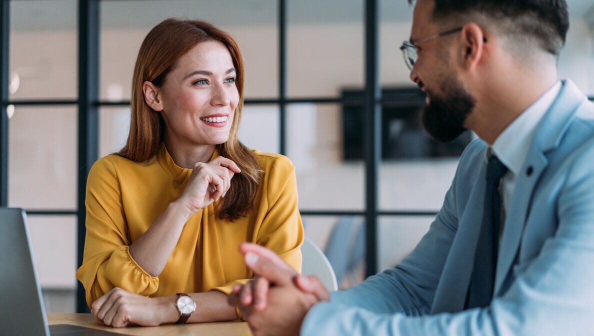 Eine Frau und ein Mann in eleganter Kleidung führen ein freundliches Gespräch an einem Tisch in einem Büro.