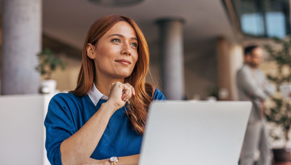 Eine Frau im blauen Pullover sitzt vor einem Laptop und schaut zufrieden lächelnd in ein Büro.