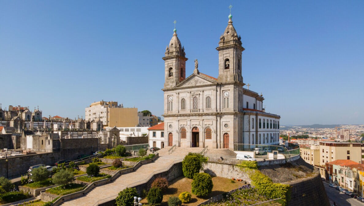 Kirche mit Zwillingstürmen auf einem Hügel über den Dächern einer südländischen Großstadt unter blauem Himmel.