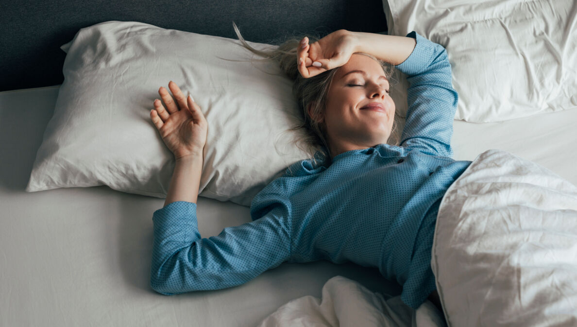 Eine zufrieden lächelnde Frau im blauen Pyjama liegt mit geschlossenen Augen in einem Bett mit weißer Bettwäsche bei Tageslicht.