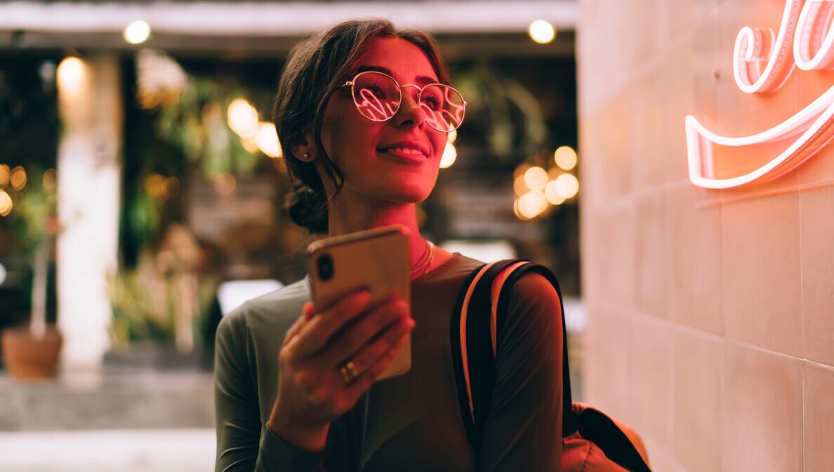 Eine junge Frau hält lächelnd ein Smartphone vor sich in der Hand, in ihrer Brille spiegelt sich der Schriftzug eines roten Logos an einer Wand.