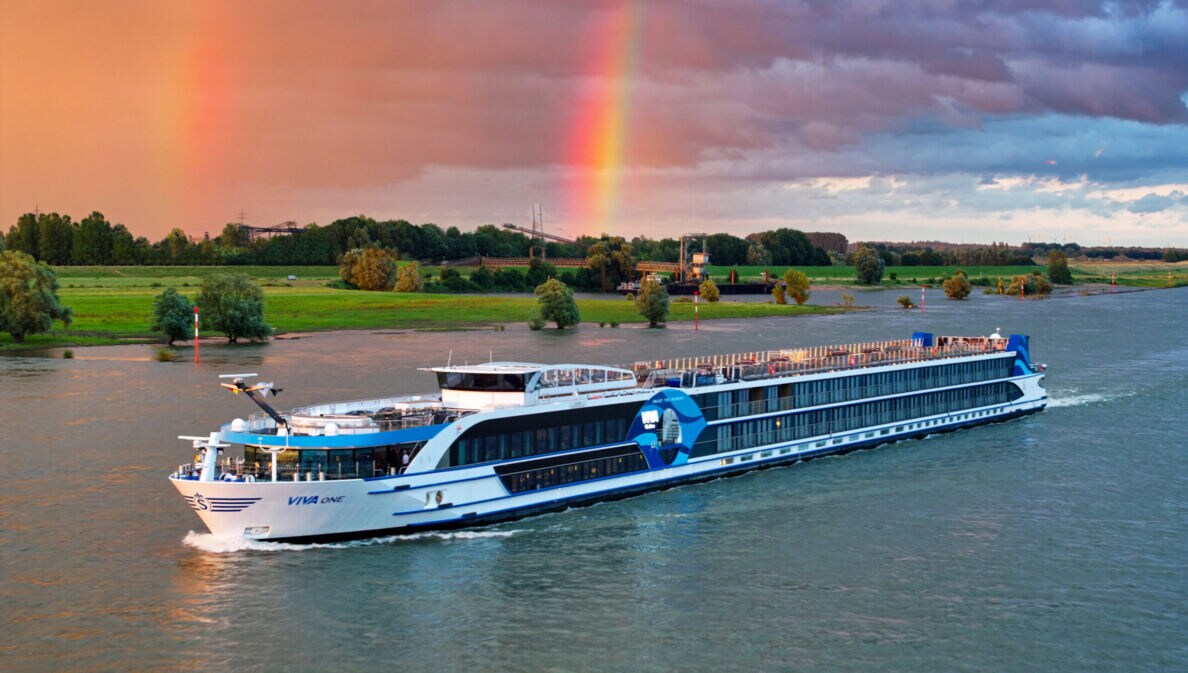 Flusskreuzfahrtschiff Viva One fährt vor grünem Flussufer mit Regenbogen am bewölkten Himmel.