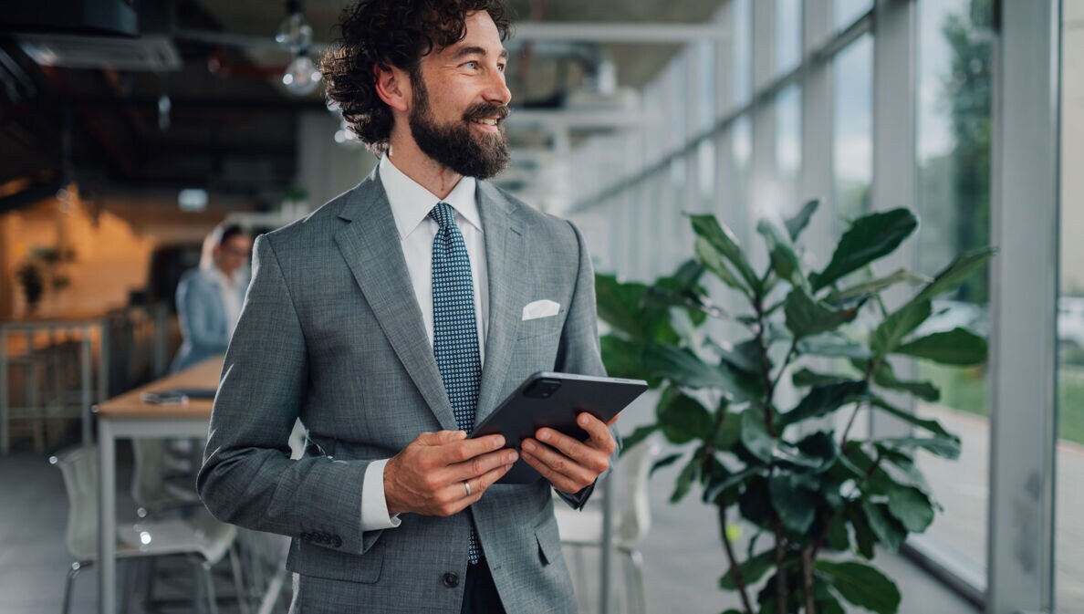 Halbporträt eines lächelnden Mannes im grauen Anzug mit Einstecktuch und Krawatte, der mit einem Tablet in einem Büro steht.