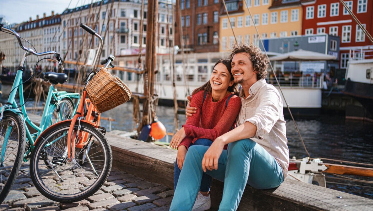Ein lachendes Paar sitzt eng beieinander auf einer Kaimauer an einem Kanal mit bunten Häusern im Stadtzentrum bei Sonnenschein, neben ihnen stehen zwei Fahrräder auf Kopfsteinpflaster.