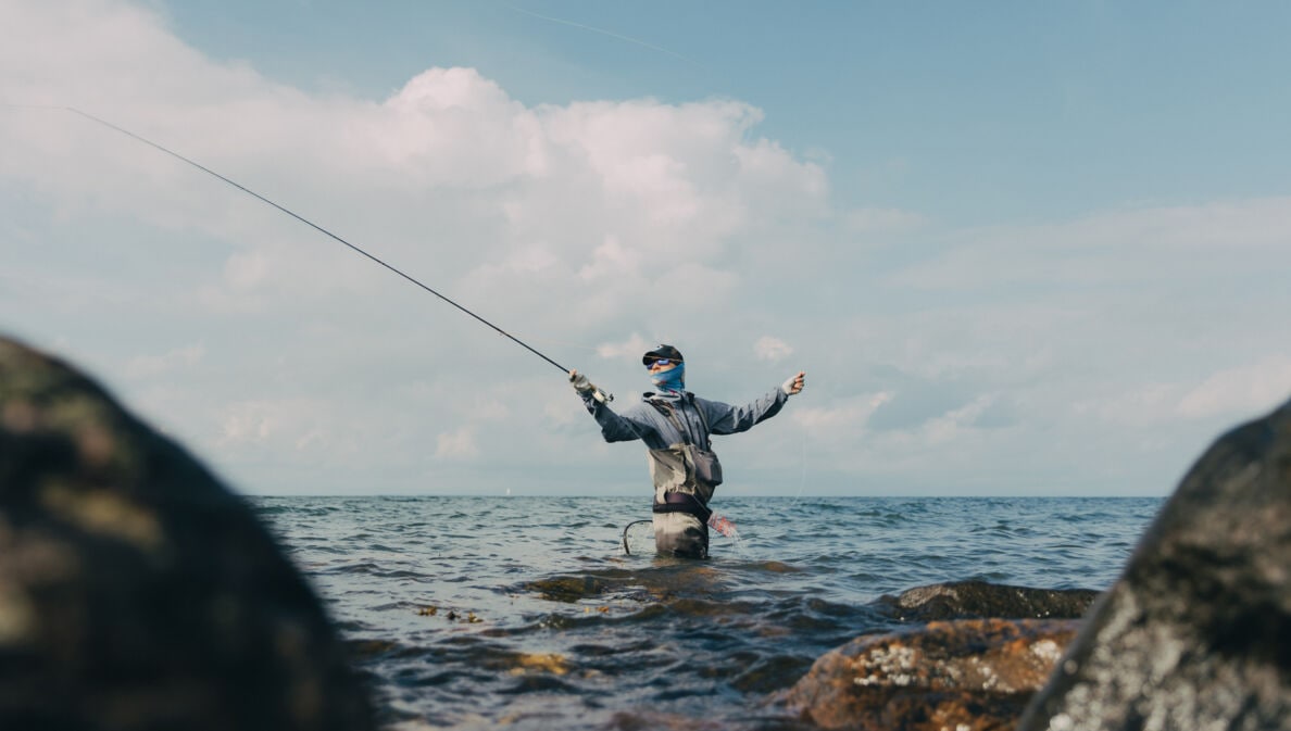 Angler steht hüfttief im Wasser zwischen Felsen und wirft eine Angelrute aus, bewölkter Himmel im Hintergrund.