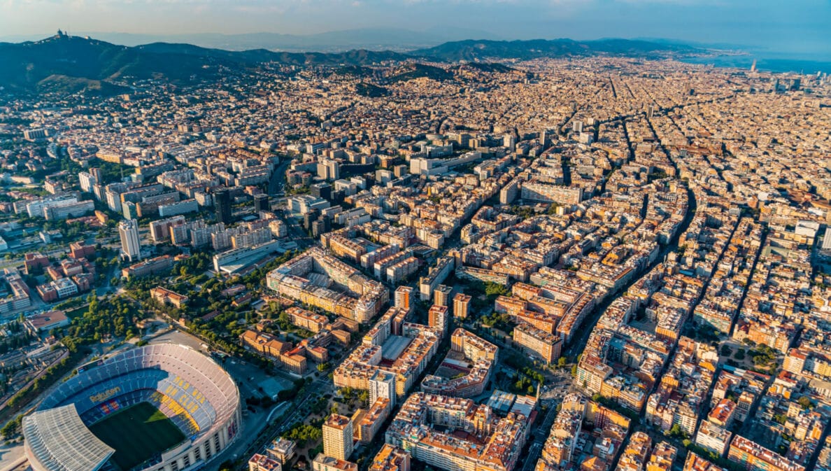 Stadtpanorama von Barcelona mit Fußballstadion aus der Luftperspektive, im Hintergrund eine Hügelkette und Meer.