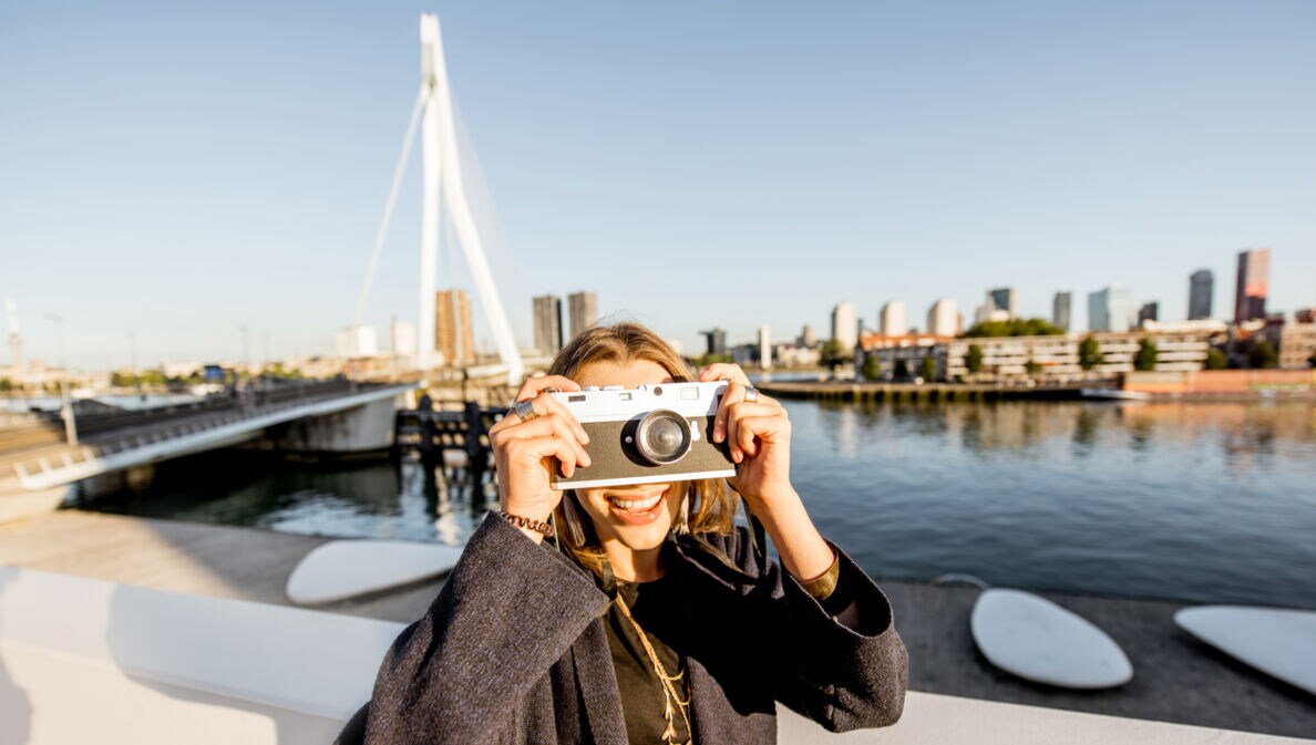 erson mit dunkelgrauem Mantel hält eine weiße Sofortbildkamera vor das Gesicht, im Hintergrund die Skyline von Rotterdam und eine Brücke über Wasser.