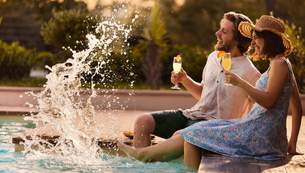 Eine Frau und ein Mann sitzen am Rand eines Pools mit einem Getränk in der Hand und spritzen mit ihren Füßen.