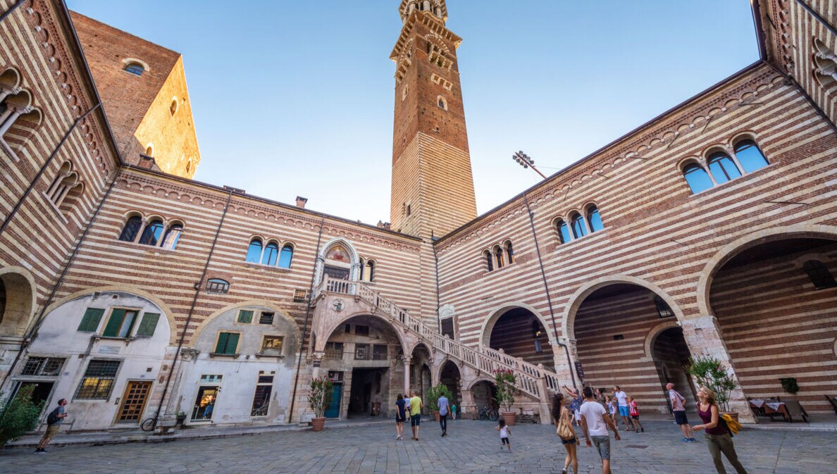Personen in einem italienischen Palastinnenhof mit gestreifter Fassade und Außentreppe unter einem Turm vor blauem Himmel.