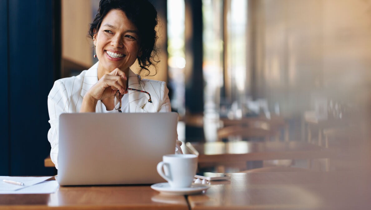 Eine Frau sitzt zufrieden lächelnd an einem Laptop an einem Tisch mit Kaffeetasse in einem Café.