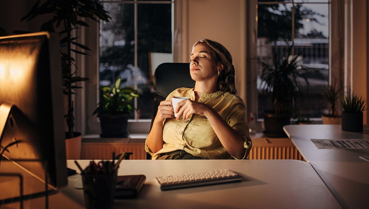Eine Frau mit einer Tasse in den Händen lehnt mit geschlossenen Augen in einem Schreibtischstuhl an einem Schreibtisch am Abend.