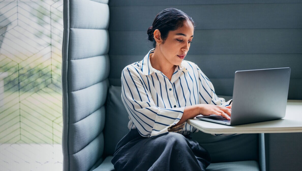 Eine Frau mit Laptop vor sich auf dem Tisch in einer gepolsterten Sitzecke.