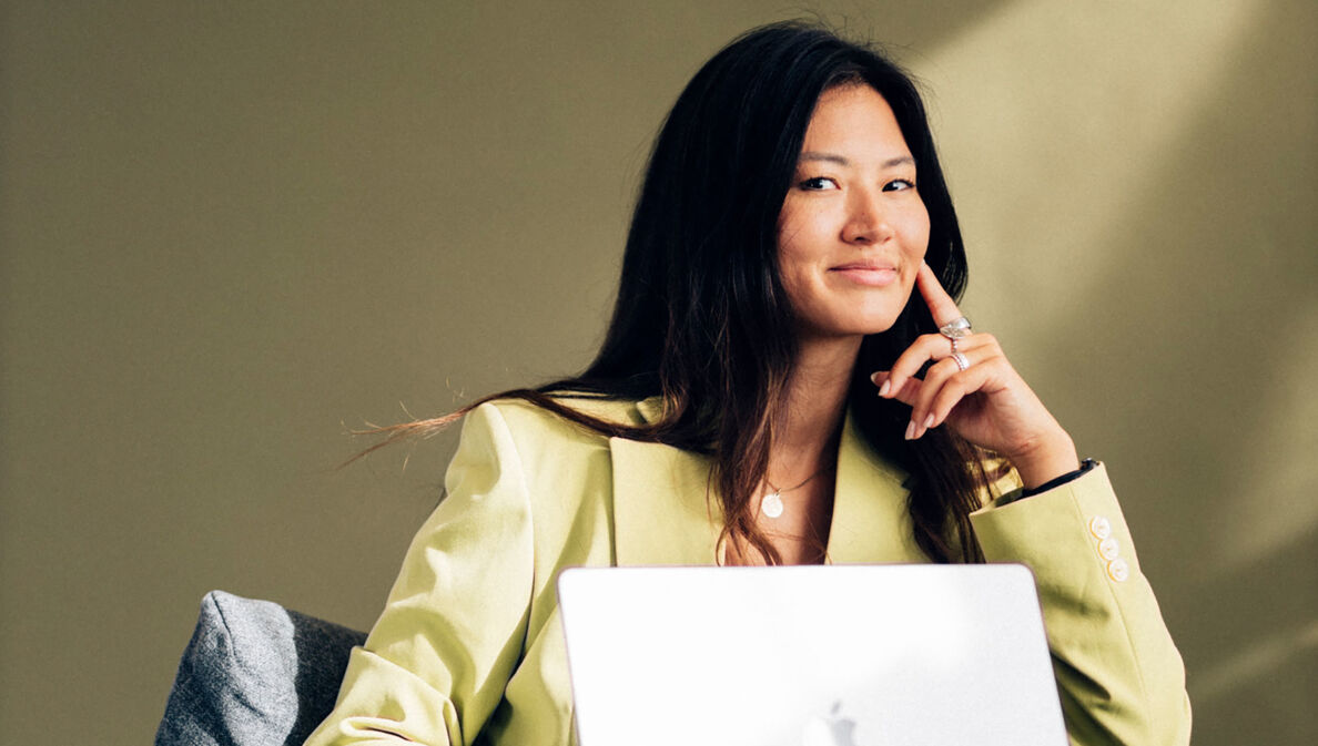 Eine Frau in einem hellgelben Blazer mit aufgestütztem Arm und einem aufgeklapptem Laptop in einem Sessel lächelt dezent in die Kamera.