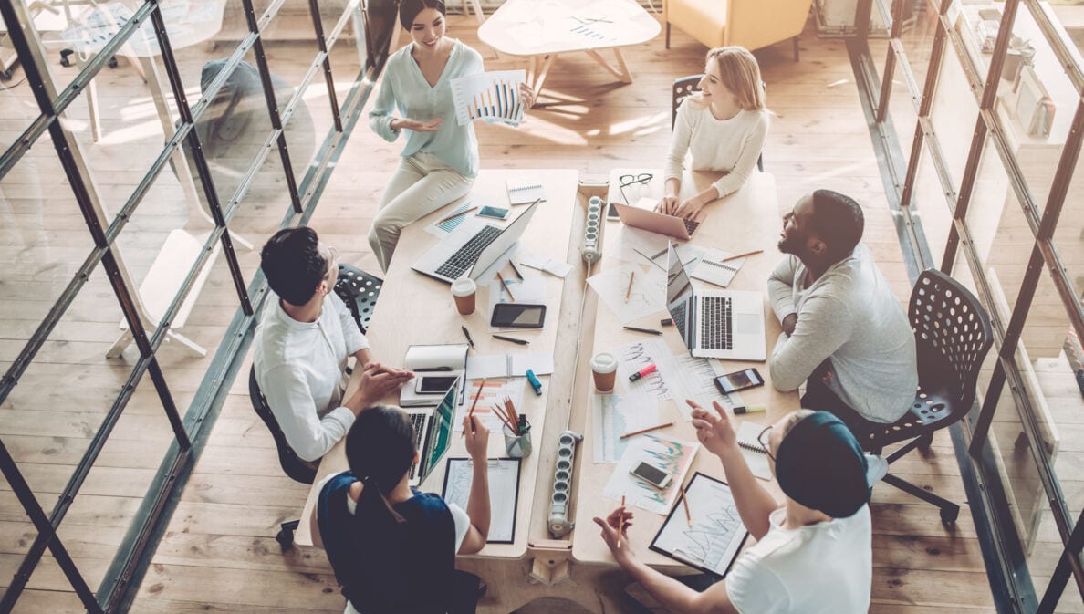 Vogelperspektive in einem Büro mit großem Tisch, an dem mehrere Personen mit Laptop sitzen und miteinander sprechen.