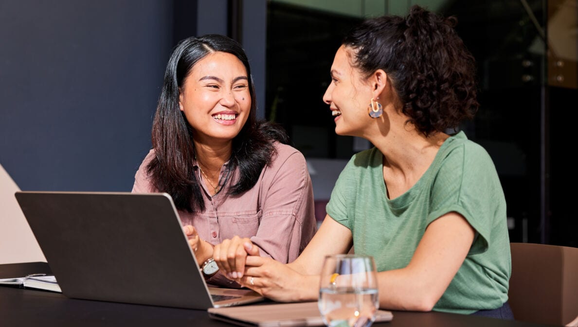 Zwei sich zugewandte lachende Frauen sitzen nebeneinander an einem Tisch, vor ihnen steht ein Laptop.