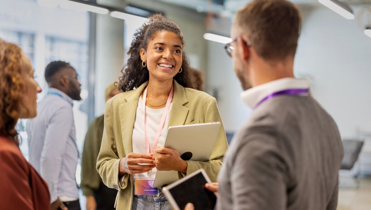 Eine junge Frau im Blazer mit Laptop in der Hand steht in einem Konferenzraum und unterhält sich mit zwei weiteren Personen.