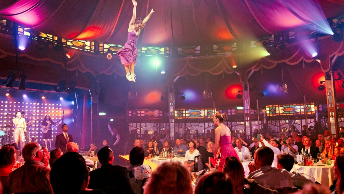 Personen sitzen beim Dinner an Tischen in einem Spiegelzelt während einer Show mit Akrobatinnen.