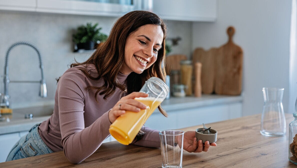 Eine lächelnde Frau gießt ein gelbes Getränk in eine Glas an einer Küchentheke.