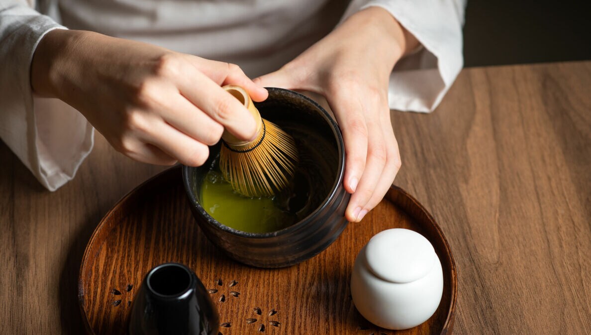 Nahaufnahme von zwei Händen bei der Zubereitung eines grünen Tees mit einem Bambusbesen in einer dunklen Keramikschale auf einem Holztisch.