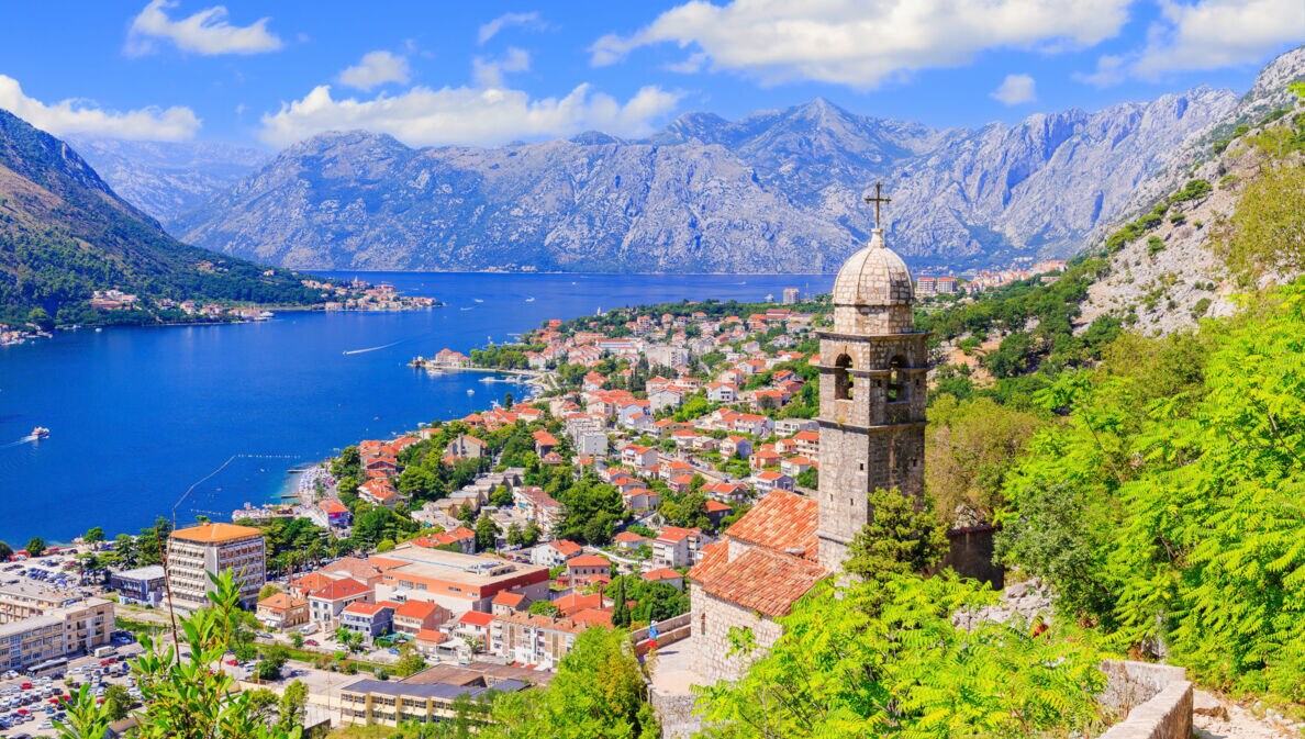 Blick auf die Stadt Kotor mit Häusern und Kirchturm, im Hintergrund Berge und eine Bucht.