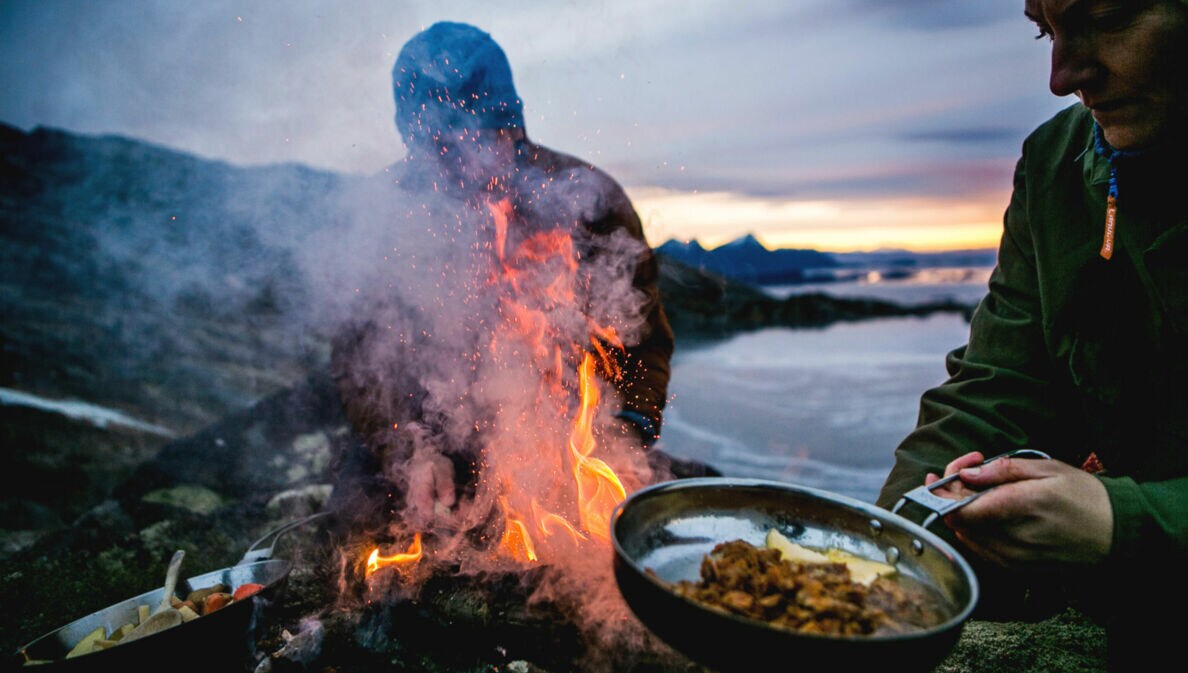 Zwei Personen bereiten Speisen in Pfannen über einem Feuer an einer schroffen Felsküste zu bei Abenddämmerung.