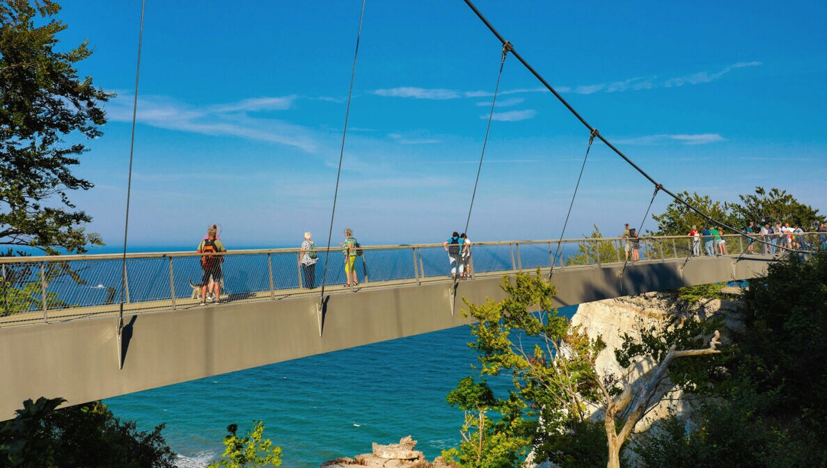 Mehrere Personen laufen auf einer schmalen Brücke zwischen Kreidefelsen am Meer unter blauem Himmel bei Sonnenschein.