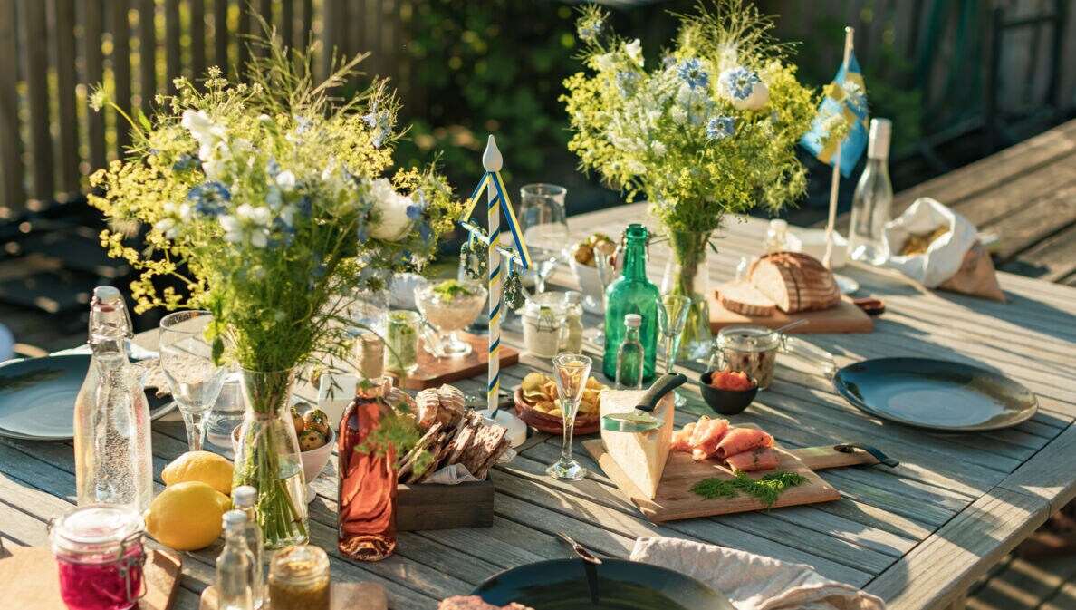 Gedeckter Holztisch für ein Mittsommer-Essen im Freien, mit Brot, gebeiztem Lachs, Käse, Eingelegtem, Getränken und frischen Blumensträußen.