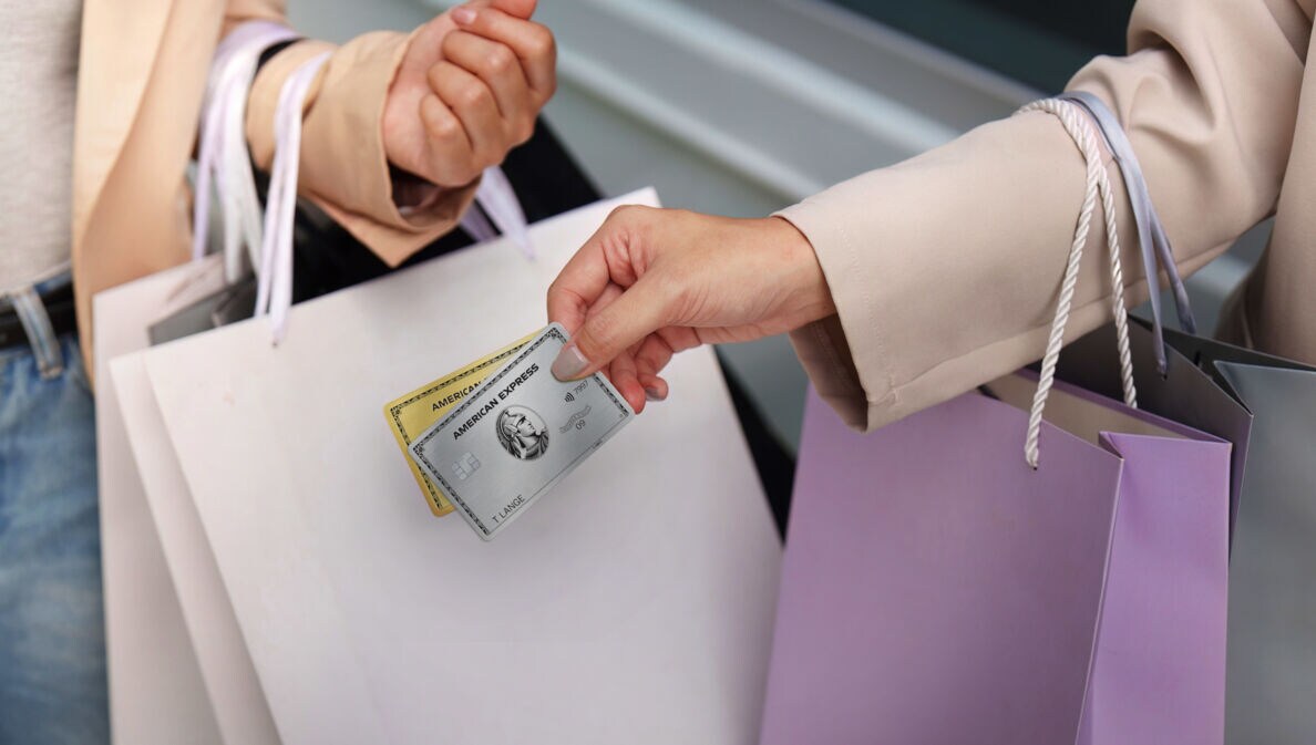 Nahaufnahme von zwei Armen mit Einkaufstüten, eine Hand hält eine goldene und eine silberne Kreditkarte von American Express.