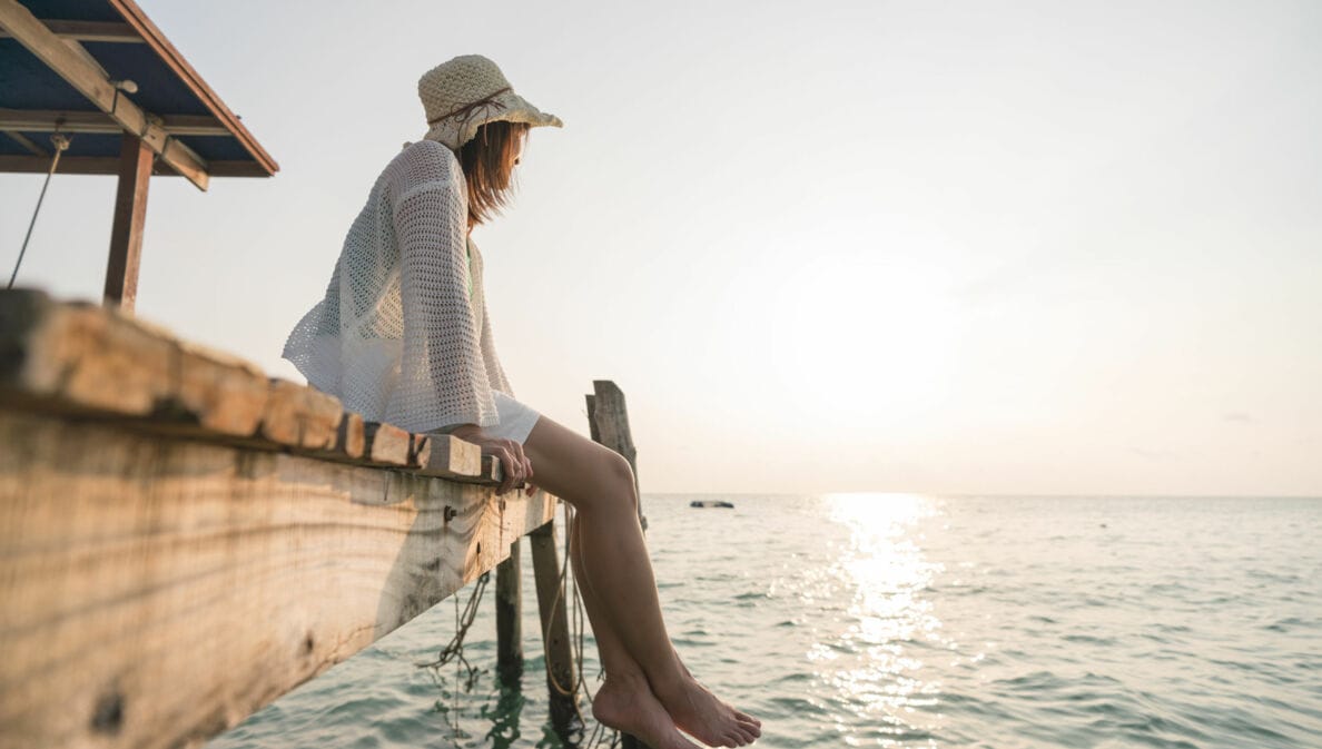 Frau mit Sonnenhut und barfuß, die auf einem Holzsteg sitzt und aufs Wasser schaut.