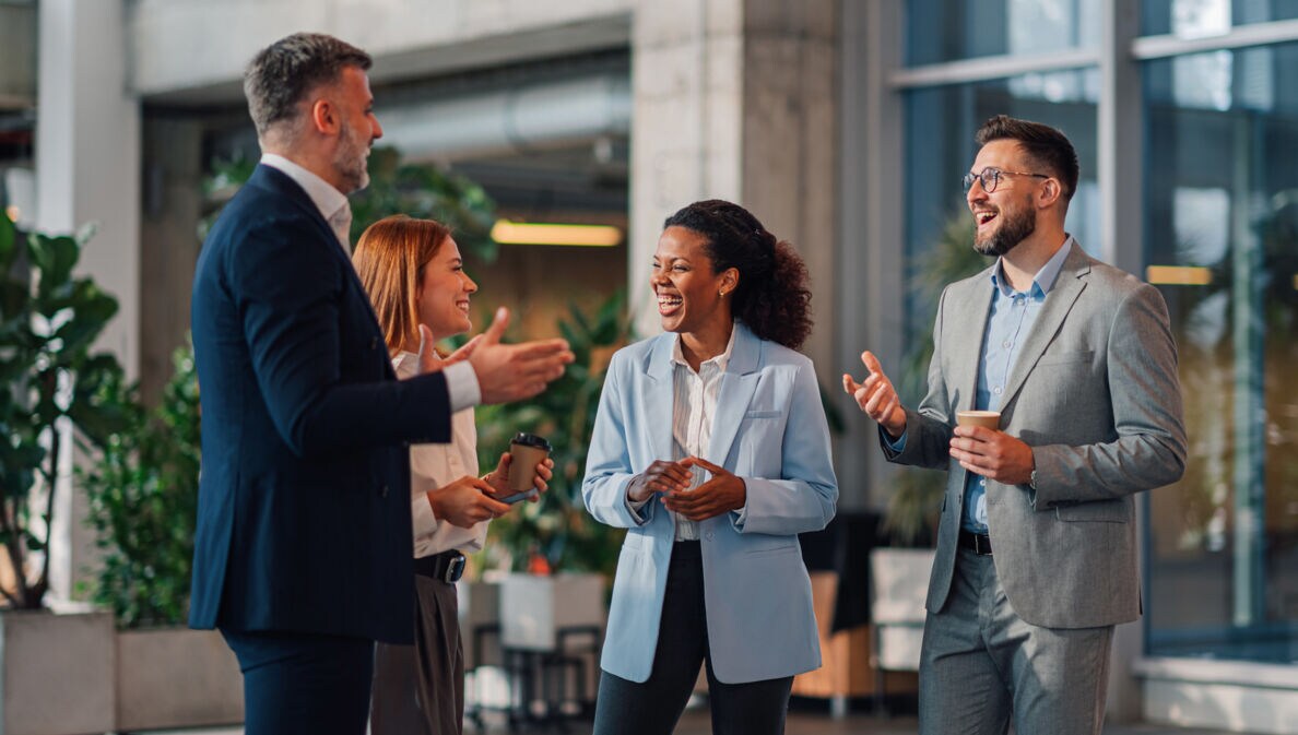 Zwei Frauen und zwei Männer in Business-Kleidung stehen beieinander und kommunizieren lächelnd in einem Foyer.